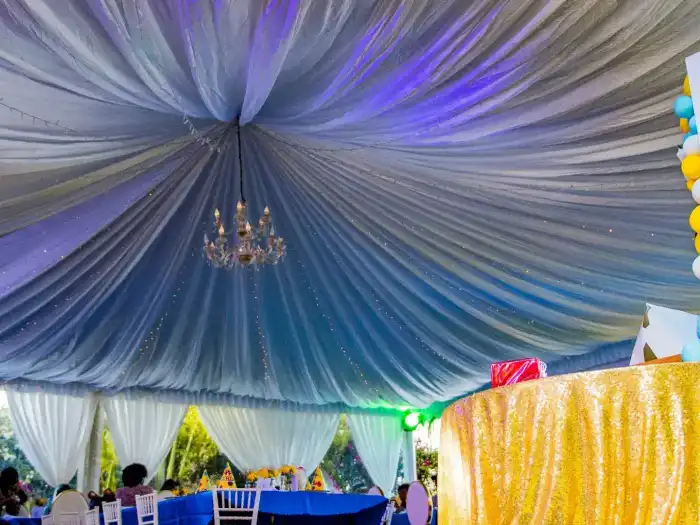 Tent interior with blue lighting, white ceiling drapes, a crystal chandelier, and gold sequin