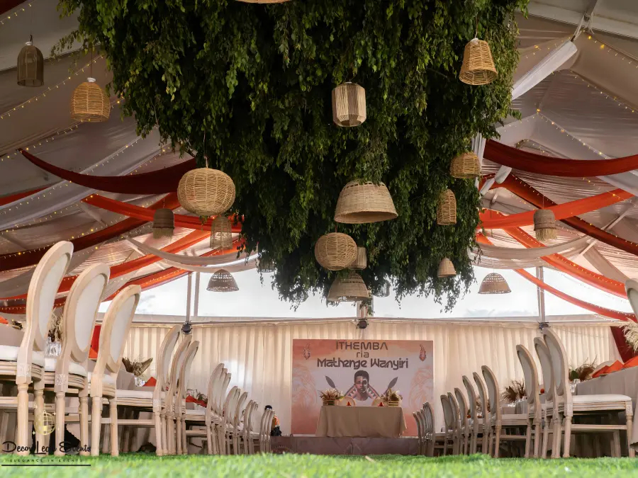 Elaborate ceiling decor inside a tent featuring rust and white drapes, and a large central greenery chandelier with woven pendant lights.