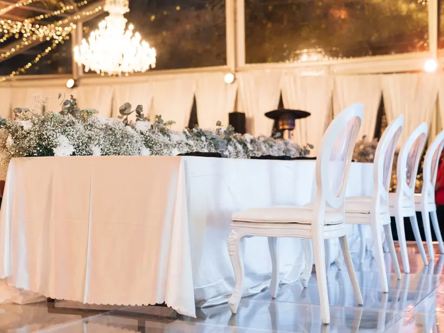 Formal white head table with a lush baby's breath runner and white Louis chairs, situated on a glossy floor under a crystal chandelier.