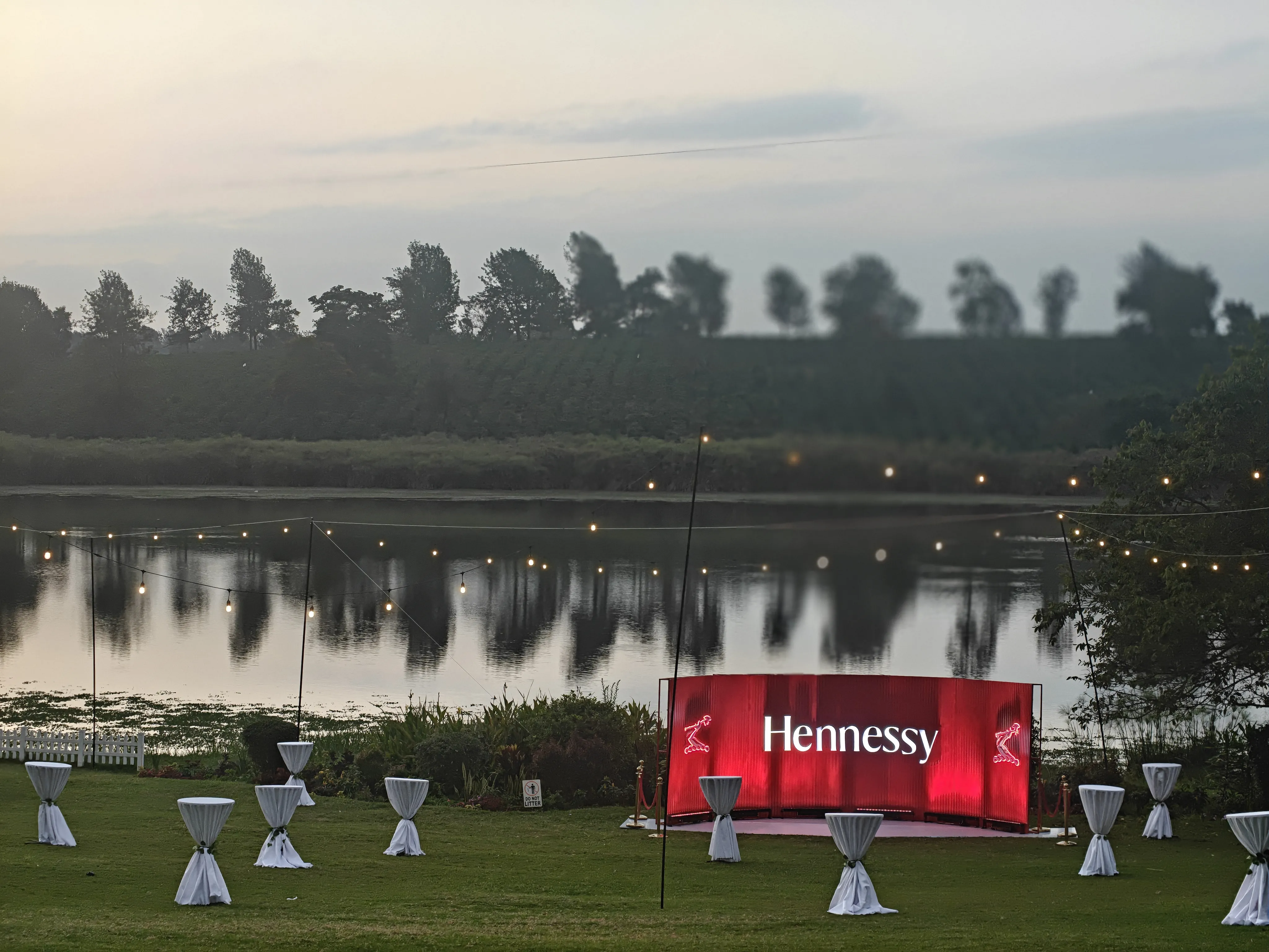 Outdoor cocktail hour setup by a lake with white-covered high tables, string lights, and a red "Hennessy" bar backdrop.