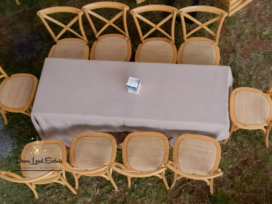 Overhead view of a rectangular banquet table with beige linen and eight wooden cross-back chairs with rattan seats on a lawn.