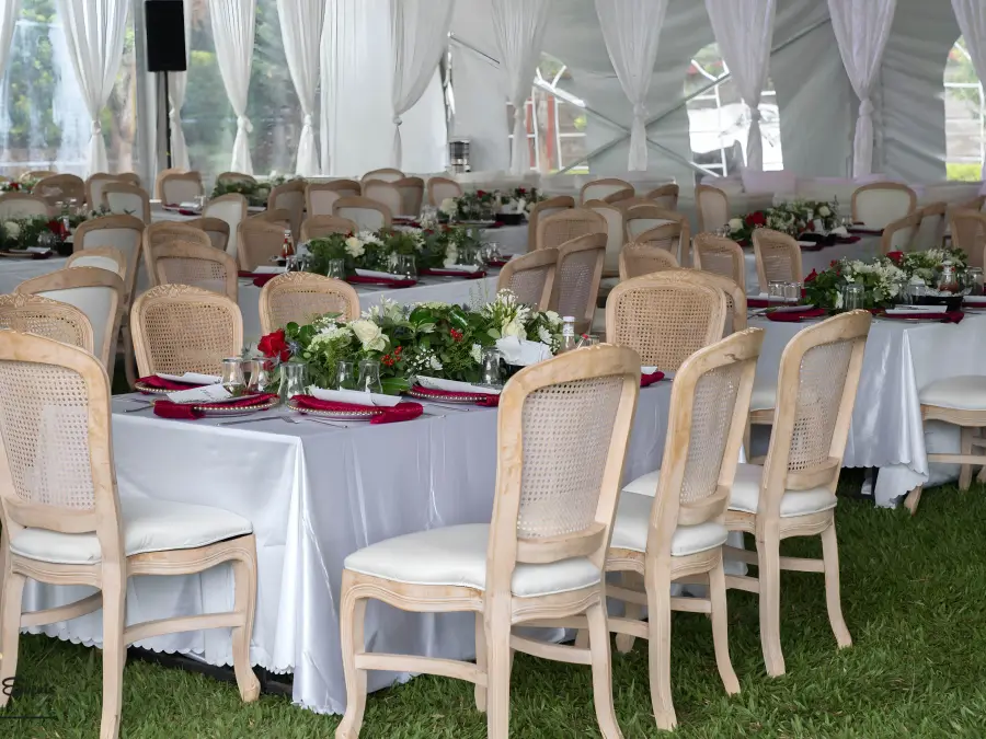 Interior reception setup with rectangular tables, white cloths, maroon runners, and wooden Louis chairs with cane backing.