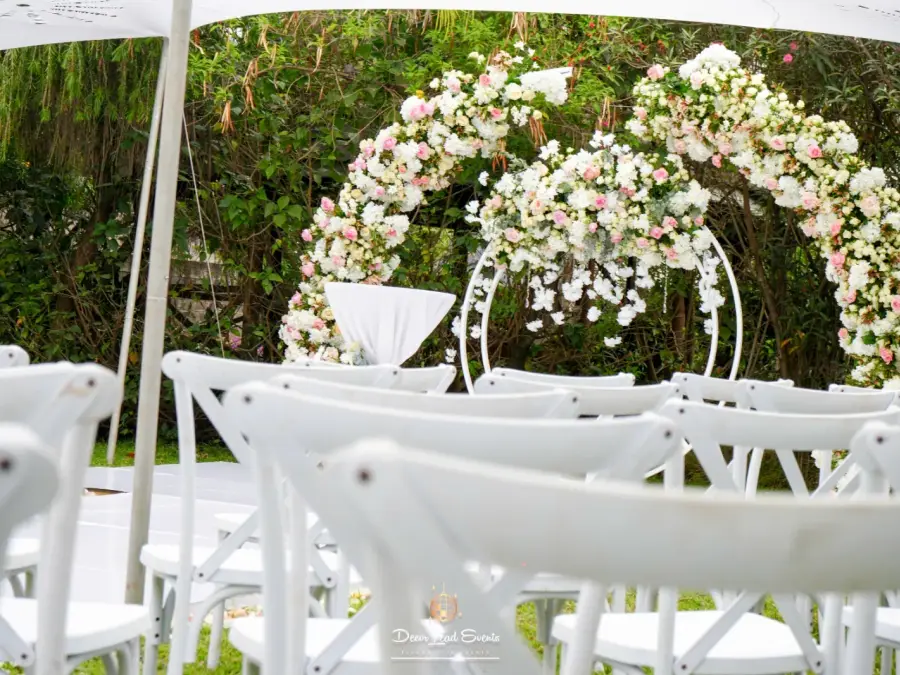 Garden wedding aisle with a red carpet leading to a floral arch, flanked by white cross-back chairs on a green lawn.
