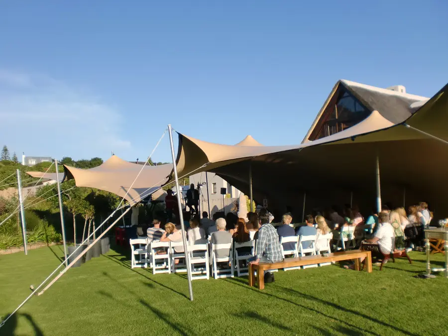 Daytime wide shot of a beige stretch tent set up on a lawn, with guests seated on white folding chairs facing a small stage.