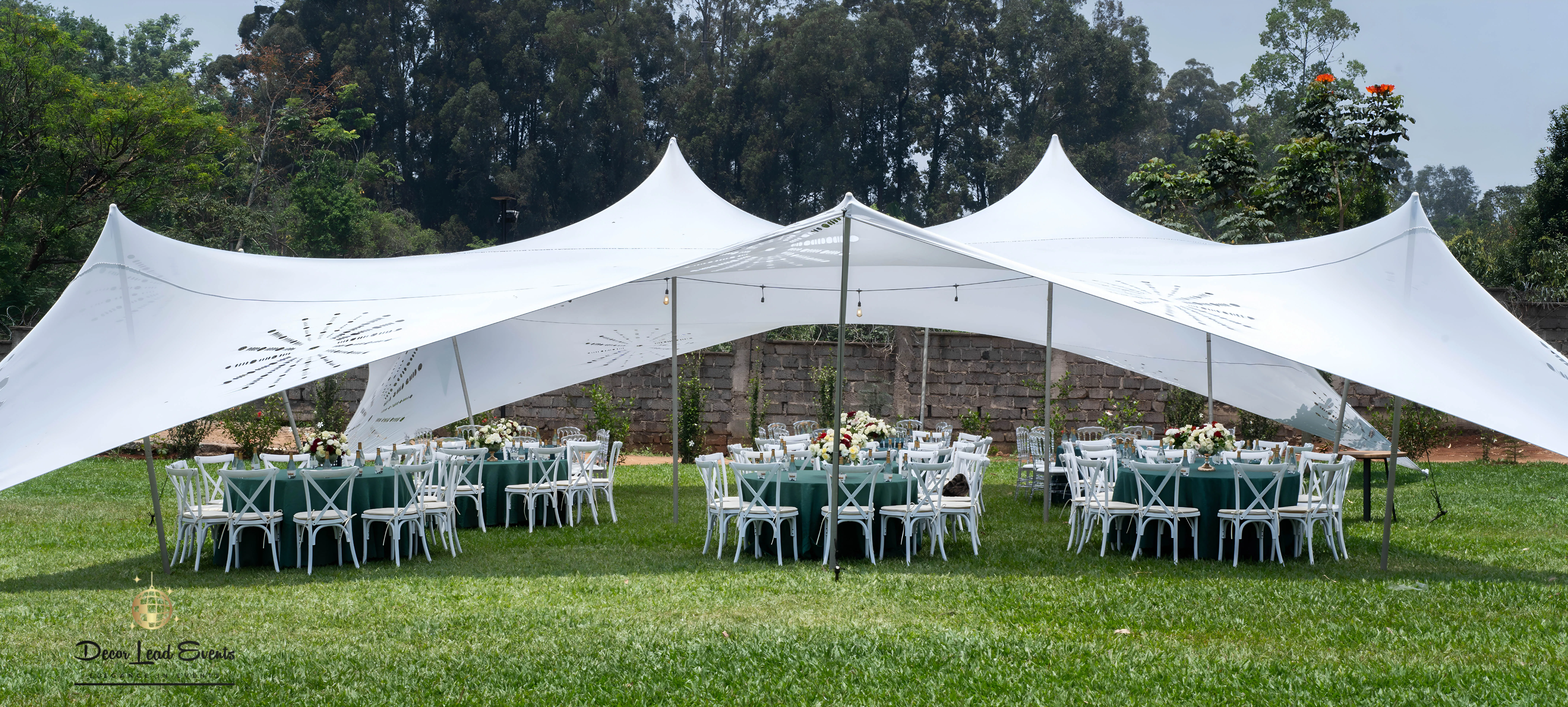 Large white stretch tent with decorative cutouts set up on a green lawn, covering round tables with green linens and white cross-back chairs.