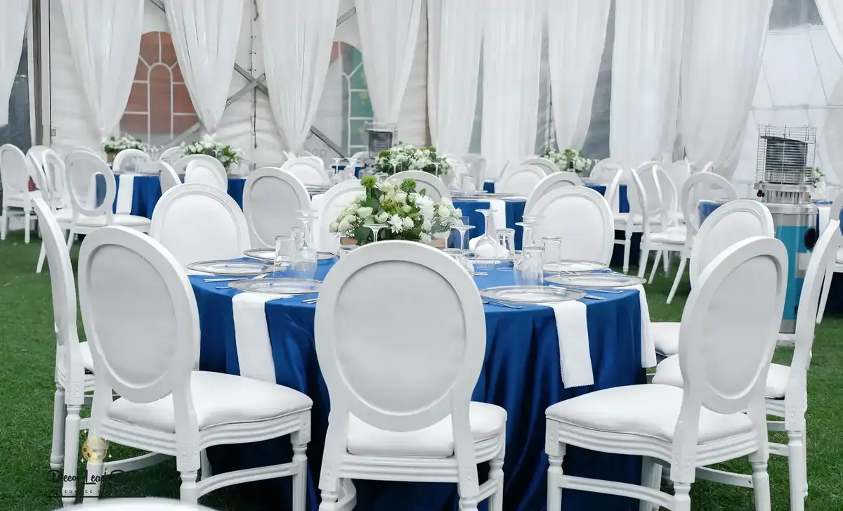 Wide interior shot of a white event tent with draped ceilings, blue round tables, white Louis